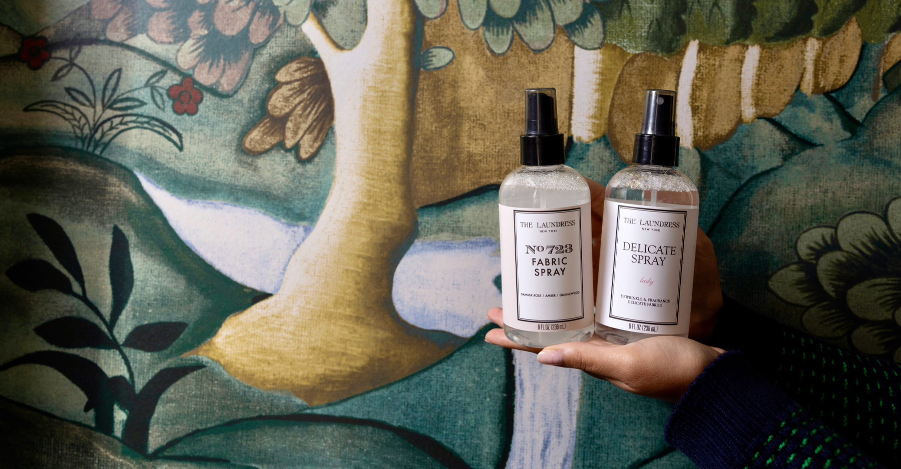 The Laundress No. 723 Fabric Spray and Delicate Spray in the scent Lady are held in front of a serene landscape mural.